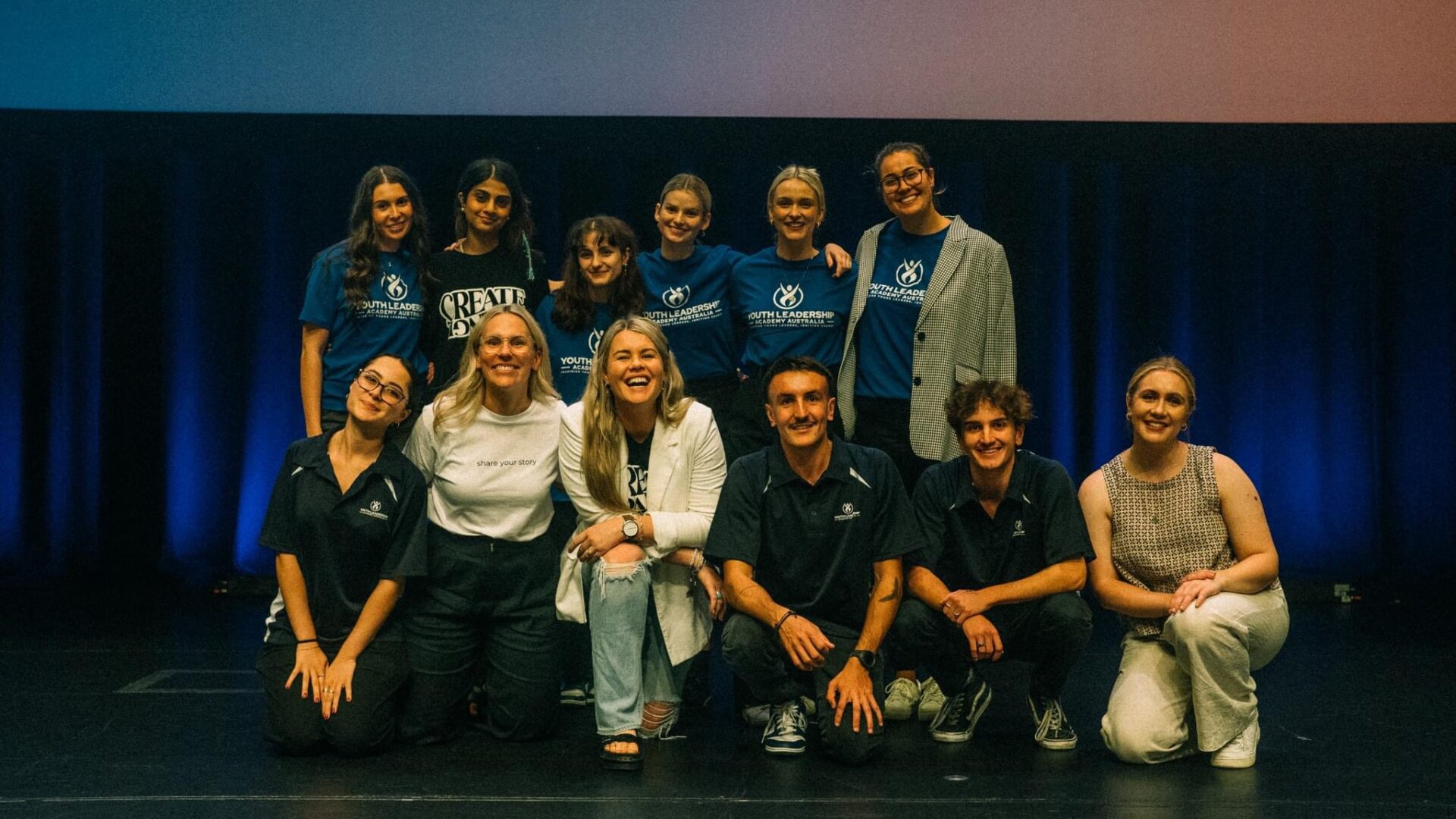 Image NA group of young adults and facilitators from the Youth Leadership Academy Australia YLAA pose together on stage smiling proudly after an event Some wear branded YLAA shirts in blue while others wear casual or business-casual attire The backdrop features dark curtains with soft blue lighting creating a professional yet welcoming atmosphere The group stands and kneels in two rows exuding confidence unity and leadershipot Found
