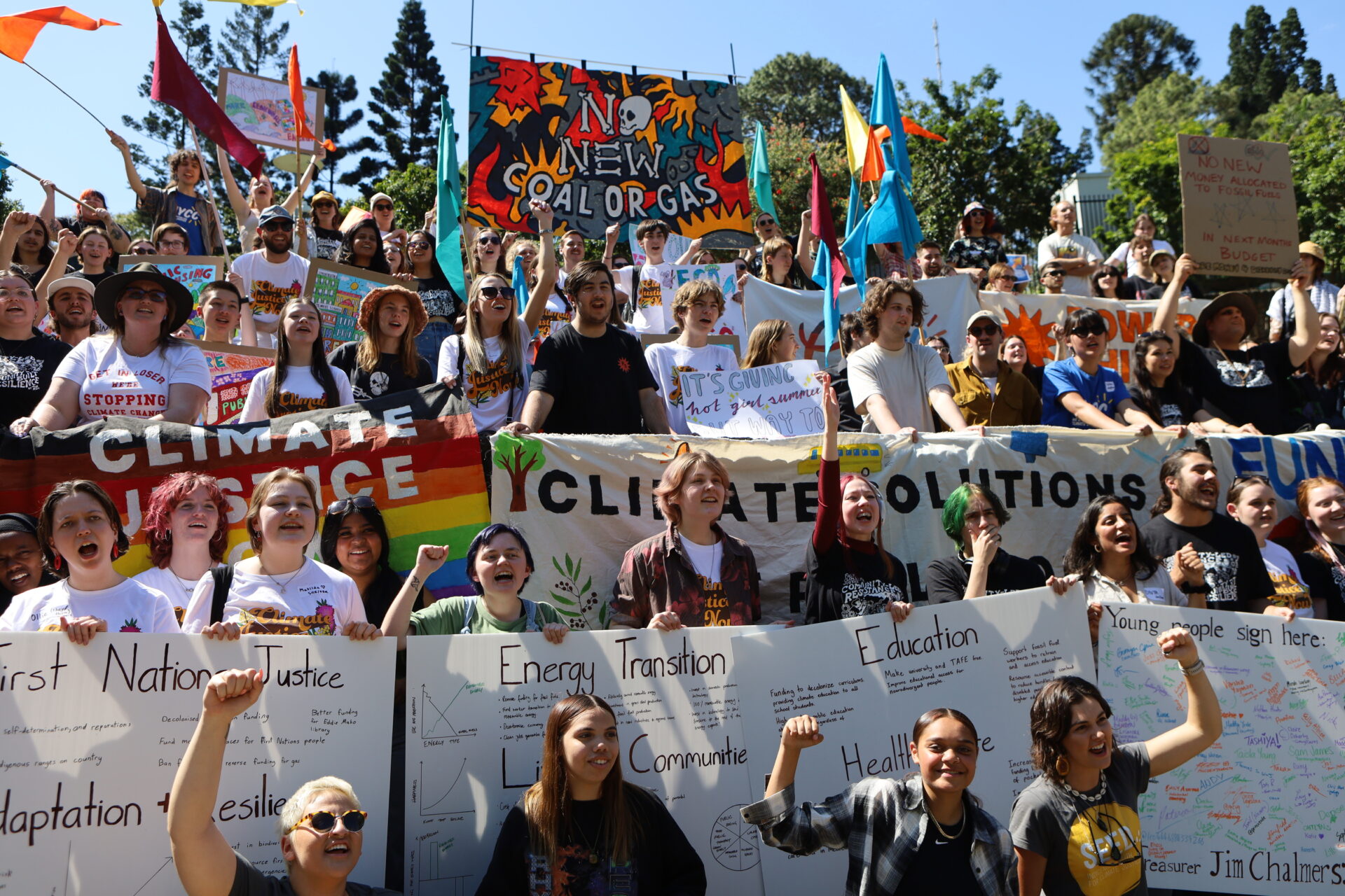 IA large diverse group of young people gather outdoors at a climate protest holding colourful banners and handmade signs calling for climate justice energy transition and an end to fossil fuels Prominent messages include No New Coal or Gas Climate Justice and Energy Transition Many participants are smiling cheering and raising their fists in solidarity The atmosphere is energetic and hopeful with vibrant flags and trees in the backgroundmage Not Found